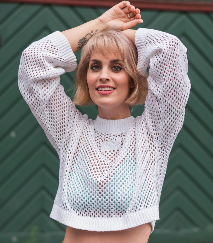 Porträt von Alina Arenz, Künstlerin und Artistin im Ensemble des Kunz Theatre, lächelnd in einem weißen Netzpullover vor grünem Hintergrund.