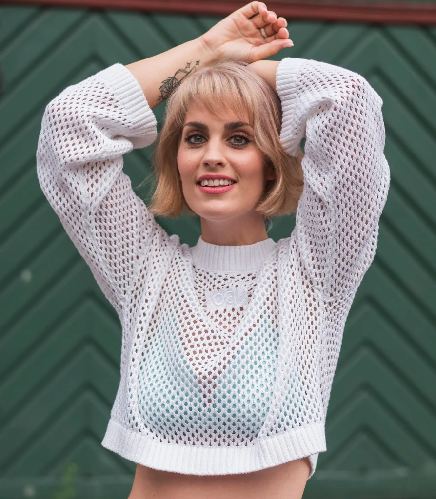 Porträt von Alina Arenz, Künstlerin und Artistin im Ensemble des Kunz Theatre, lächelnd in einem weißen Netzpullover vor grünem Hintergrund.